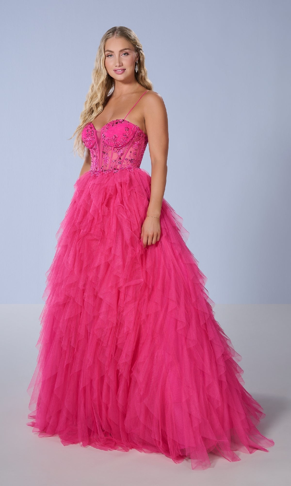 A woman models the Tiffany 16149 long prom dress, featuring a bright pink, beaded fitted bodice and a voluminous tulle skirt with cascading ruffles, set against a plain light background.